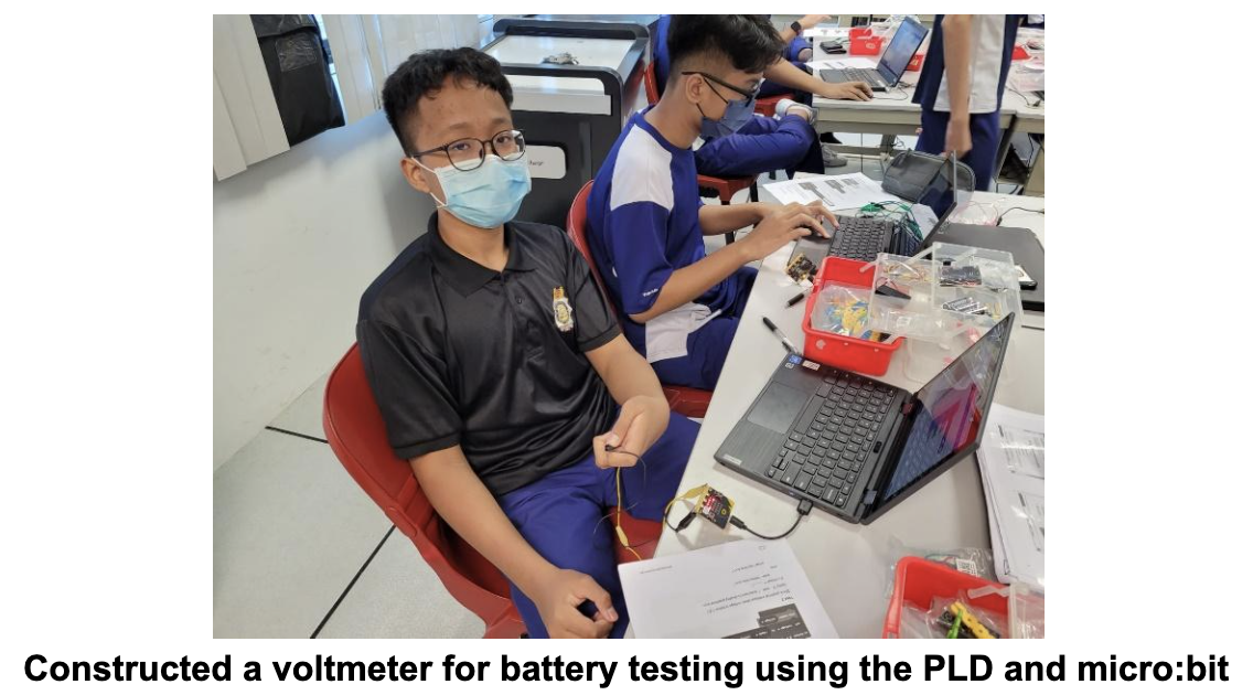 Two masked students sit at a desk with laptops and electronics components during a classroom activity.